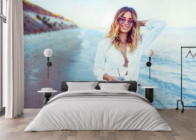 Pretty blonde woman in the shape of a heart sunglasses, white shirt and stripped swimsuit relaxing on beach and drinks coctail. summer vacation concept. Wall mural