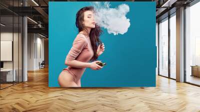 Smiling hot woman in bodysuit standing and vaping on blue background. Wall mural