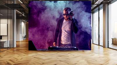 Young stylish man in glasses posing behind mixing console on colored smoke studio background. Wall mural