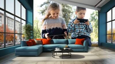 Children use gadgets while sitting in the park outdoors, games on smartphone and tablet Wall mural