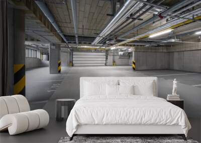 A spacious underground parking garage with exposed pipes on the ceiling, yellow and black striped pillars, concrete walls, and marked parking spaces, with no cars or people present. Wall mural