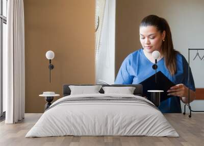 Young hispanic nurse reading a clinical report in a hospital hallway, wearing blue uniform. Wall mural