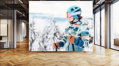 Woman in skiing clothes with helmet and ski googles on her head with ski sticks. Winter weather on the slopes. On top of a mountain and enjoying view. Alpine skier. Winter sport Wall mural