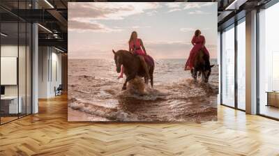 Two female riders in vibrant pink dresses leading two majestic horses along a beach coastline. Wall mural