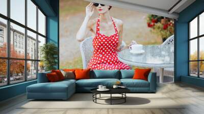 Young beautiful woman in a red dress wearing sunglasses is sitting at a table in an outdoor cafe Wall mural