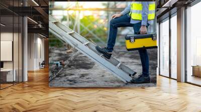 Asian maintenance worker man with safety helmet and green vest carrying tool box climbing on aluminium ladder at construction site. Civil engineering, Architecture builder and building service concept Wall mural