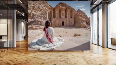 Asian woman tourist in white dress sitting and looking at Ad Deir or El Deir, the monument carved out of rock in the ancient city of Petra, Jordan. Travel UNESCO World Heritage Site in Middle East Wall mural