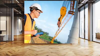 Smart Asian worker man or male civil engineer with protective safety helmet and reflective vest using digital tablet for project planning and checking architectural drawing at construction site. Wall mural