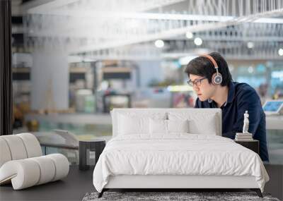 Young Asian business man listening to music by headphones while working with laptop computer in co working space. freelance or digital nomad lifestyle in urban workspace concepts Wall mural