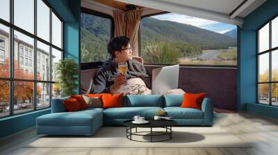 Young Asian man drinking beer and working with laptop computer on the bed in camper van with mountain scenic view through the window, digital nomad concept Wall mural