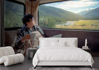 Young Asian man drinking hot coffee working with laptop computer on the bed in camper van with mountain scenic view through the window, digital nomad on road trip concept Wall mural