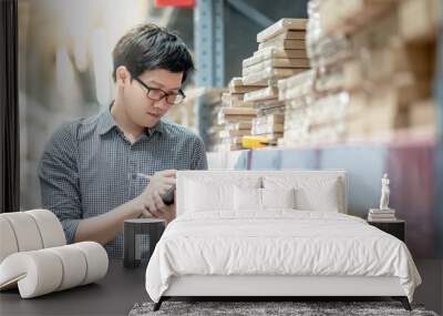 Young Asian man worker doing stocktaking of product in cardboard box on shelves in warehouse by using digital tablet and pen. Physical inventory count concept Wall mural