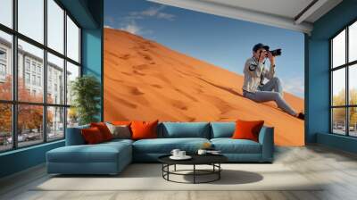 Young Asian woman traveler and photographer holding camera taking photo while sitting on sand dune in Namib desert of Namibia, Africa. Travel photography concept Wall mural