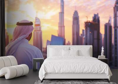 
An Arab man in a white kandura looking at a modern city skyline with skyscrapers during sunset. A business and lifestyle concept. Wall mural