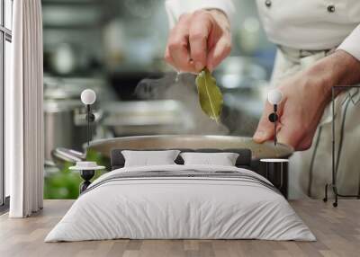 A chef places a bay leaf into a simmering soup pot, showcasing a blend of tradition and culinary expertise in a professional kitchen, with steam rising to create an inviting atmosphere. Wall mural