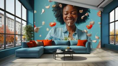 A joyful woman with curly hair smiles as garlic cloves and cherry tomatoes fly around her, adding vibrant energy to the scene. The blue background enhances the freshness of the salad she is about to Wall mural