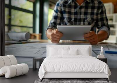 An architect in casual attire reviews blueprints on a desk using a digital tablet. The scene combines traditional paper plans with modern digital tools, emphasizing a blend of old and new methods in Wall mural