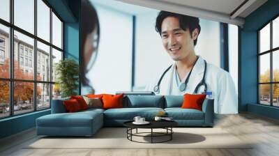 a handsome Japanese man doctor wearing white coat, smiling while having conversation with patient in clinic room. Wall mural