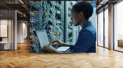 A female african american IT engineer works in a data center, analyzing network configurations on her laptop while surrounded by server racks and numerous connected cables Wall mural