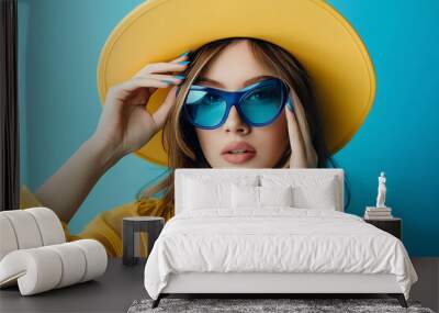 A stylish woman in a yellow hat and blue sunglasses, posing against a vibrant blue background, evokes summer vacation vibes and a carefree spirit. 
 Wall mural