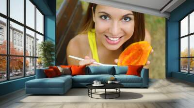 Girl eating healthy papaya in the park looking at camera. Woman eating tropical fresh fruit outdoor. Wall mural