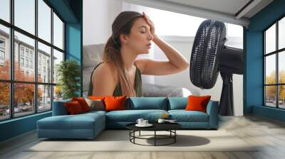 Overheated young woman in front of the working fan suffering from summer heat at home. Girl sitting in front of ventilator in hot weather. Hot summer concept. Wall mural