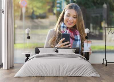 Portrait of young cheerful woman wearing coat and scarf using her phone in city street and drinking take away coffee. Wall mural