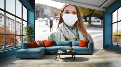 Portrait of young woman in city street wearing white surgical mask looking at camera. Girl with protective mask on face. Wall mural