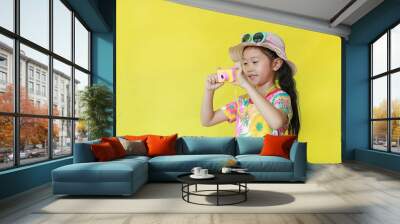 Little girl taking photo by toy camera. Child tourists in floral pattern summer dress and hat with sunglasses isolated over yellow background. Wall mural