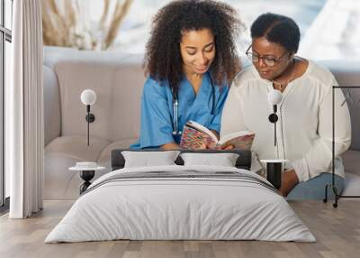 Caring nurse sitting on sofa near patient and reading book for her Wall mural