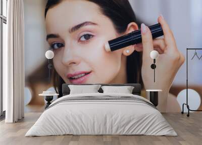 Concealer stick. Charming beautiful woman using concealer stick while putting makeup on Wall mural