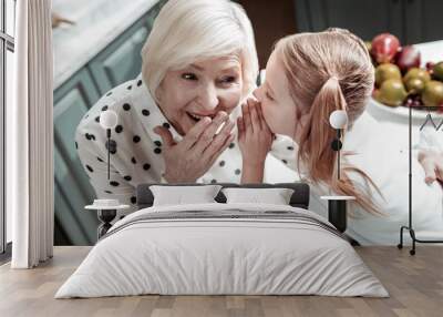 Emotional granny touching her mouth while little girl sharing secrets with her Wall mural