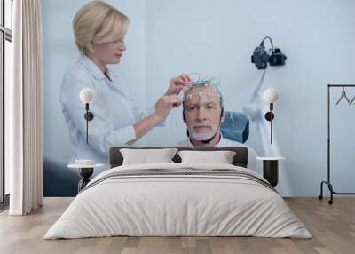 Female neurologist adjusting electrodes on gray-haired male patient head Wall mural