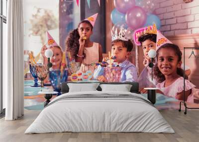 Five children. Positive relaxed pretty kids sitting together at the birthday party and blowing lovely party horns Wall mural