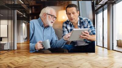 Gripping content. Loving song showing an interesting article on the tablet to his elderly father while the man holing a cup of coffee and paying attention to his son Wall mural