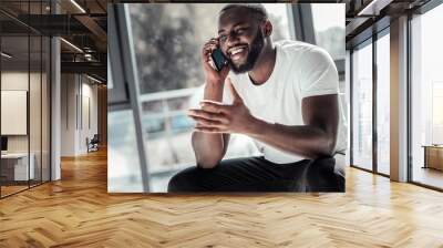 Phone conversation. Happy cheerful young man smiling and talking on the phone while sitting in the office Wall mural