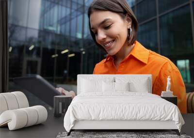 Smiling girl in orange shirt with a device looking contented Wall mural