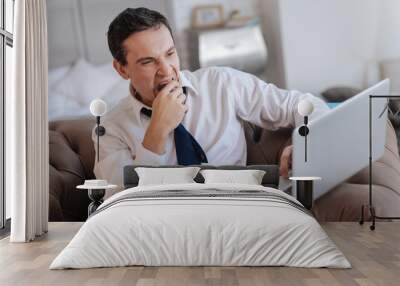 So sleepy. Tired bored sleepy man sitting in a comfortable chair and yawning while looking at the screen of his modern convenient laptop Wall mural