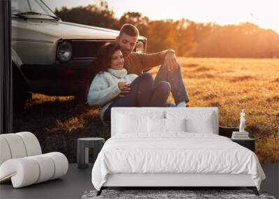 Beautiful young couple enjoying time together outdoor sitting on meadow leaning on old fashioned car embracing each other. Wall mural