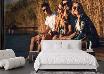 Group of friends enjoying a day at the lake. They sitting on pier talking, laughing and drinking beers. Wall mural