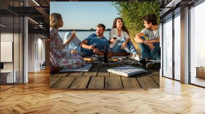 Group of friends having fun on picnic near a lake, sitting on pier eating and drinking wine. Wall mural