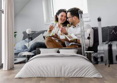 Smiling couple sitting on sofa and eating fast food salad at hotel room. Wall mural