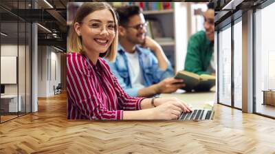 Three young students study in the school library, female student sitting in front of laptop and looking at camera. Wall mural