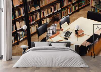 Tired young female student study in the library reading a book and using laptop for researching online. Wall mural