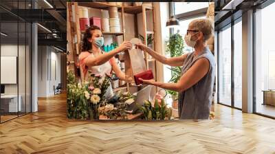 Young female florist with mask working in flower shop, selling flower arrangement to senior woman.  Wall mural