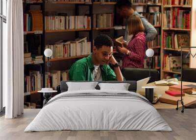 Young male student study in the library using laptop for researching online, while his friend searching the book on the bookshelf behind him. Wall mural