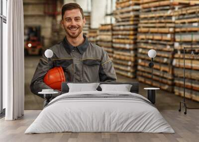 Attractive young male constructionist smiling to the camera, holding his hardhat, posing at the storage of metalworking factory. Profession, heavy industry, achievement, development concept Wall mural