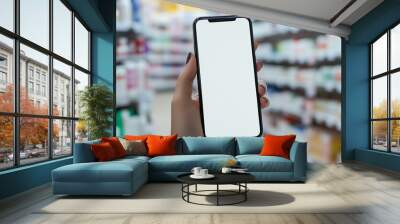 A hand holding a blank smartphone in a pharmacy aisle filled with products during daytime Wall mural