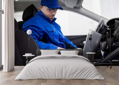 Mechanic in blue uniform working on laptop inside car to diagnose technical issues at an auto repair shop during daytime Wall mural