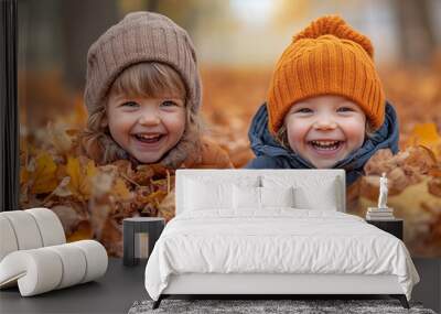 Two joyful young kids lie in a pile of colorful autumn leaves, grinning widely while wearing warm winter hats in a park filled with fall foliage Wall mural